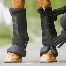 Load image into Gallery viewer, Black horse leg boots with multiple adjustable straps and protective lower cuffs, shown on a horse's legs outdoors with a person in black gloves adjusting one boot.